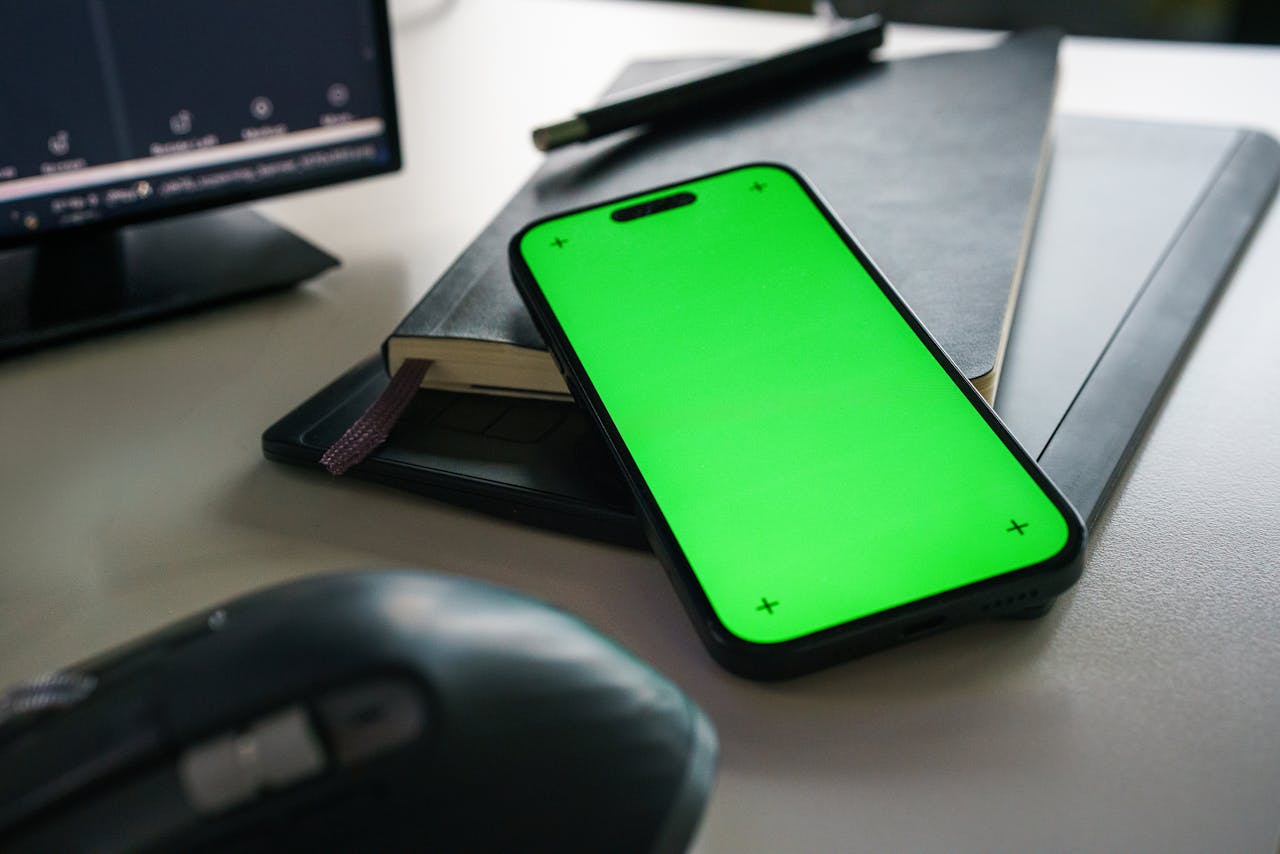 A clean and modern office desk with a smartphone displaying a green screen, notebook, and computer accessories.